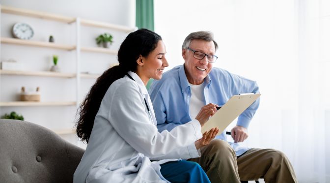 Young doctor asking senior impaired male patient in wheelchair to sign insurance policy at home. Handicapped elderly man putting his signature under surgery consent form, reading medical document
